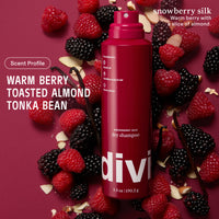 A red spray bottle of divi dry shampoo, labeled 'snowberry silk' and 'Warm Berry Toasted Almond Tonka Bean', surrounded by fresh raspberries, blackberries, and sliced almonds on a dark pink background.